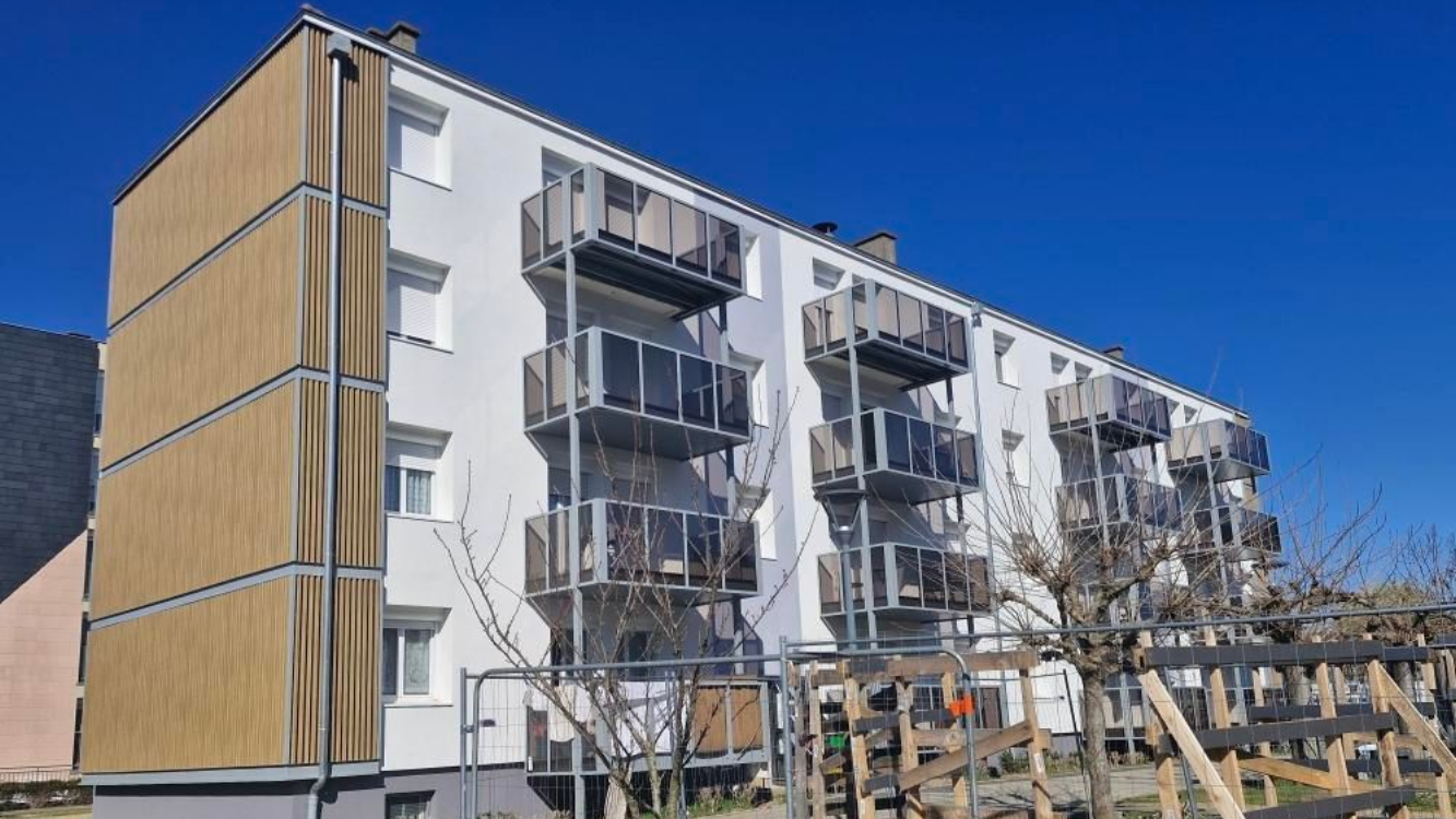 Un immeuble d'appartements blanc de quatre étages, avec des balcons en métal et un revêtement en bois sur le côté gauche, sous un ciel bleu.