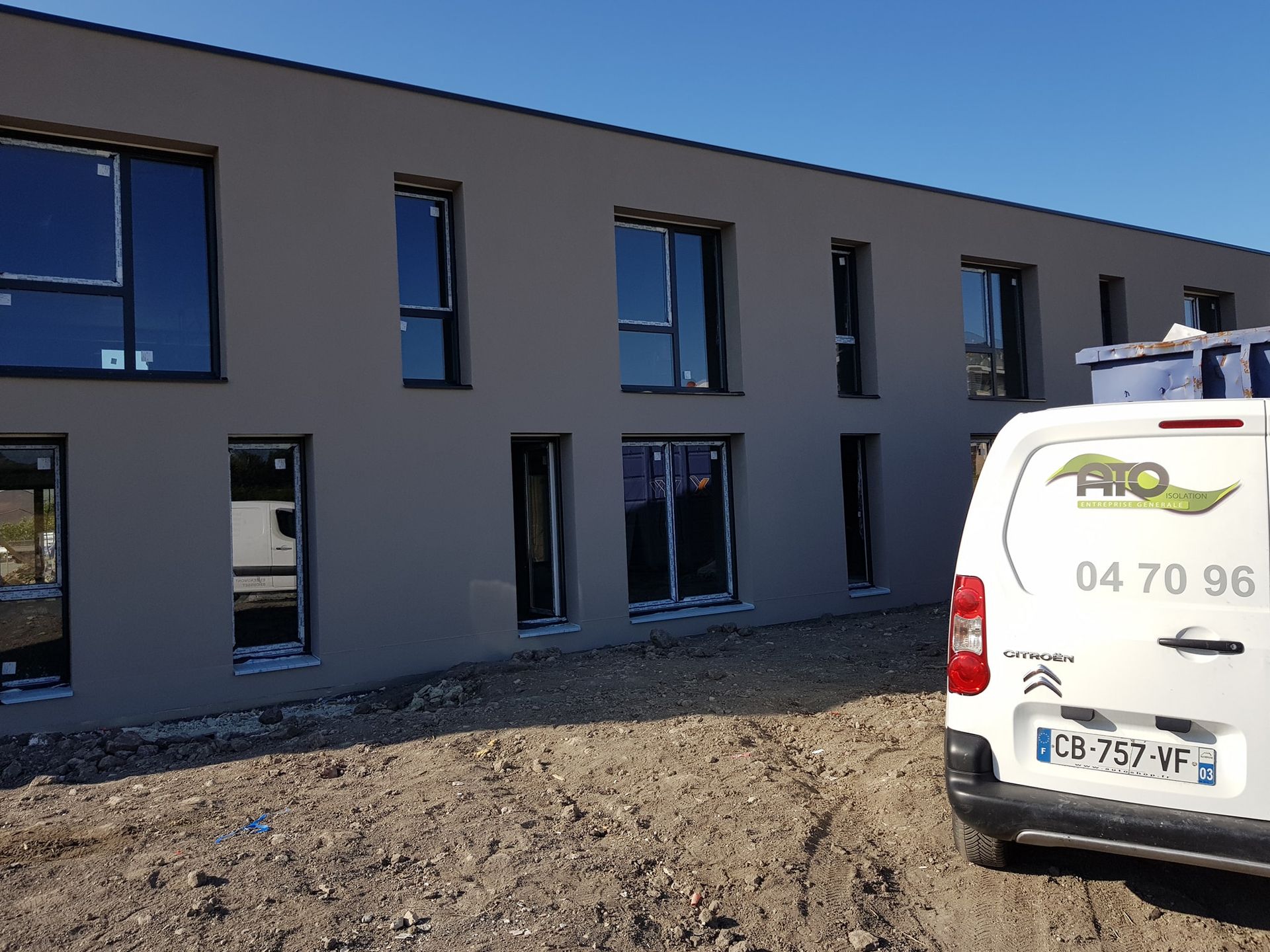 Une camionnette blanche de chantier est garée devant un bâtiment moderne de deux étages, à la façade grise et percé de plusieurs fenêtres.