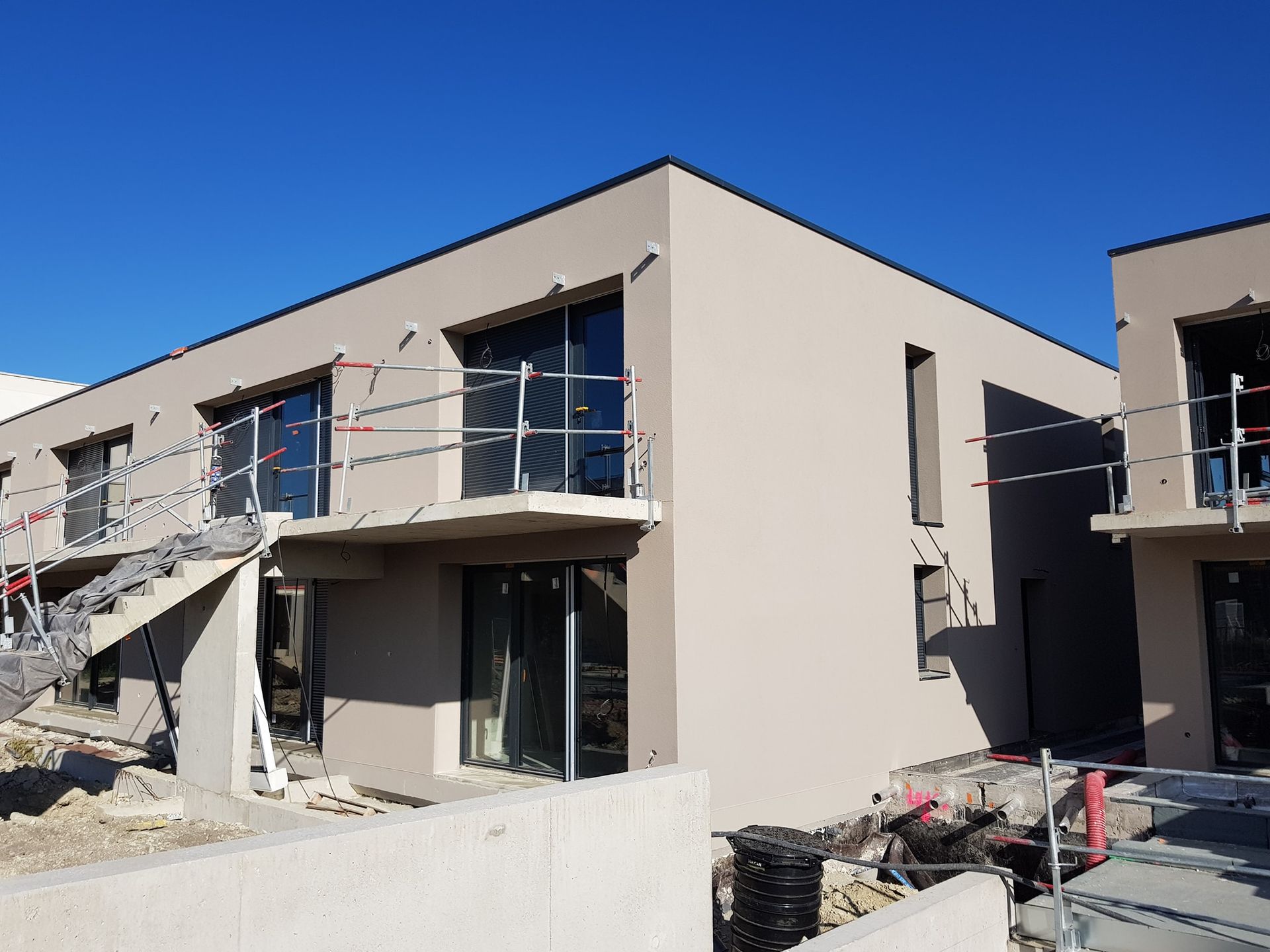 Bâtiment moderne beige de deux étages en construction, avec des garde-corps métalliques et des échafaudages, se détachant sur un ciel bleu clair.