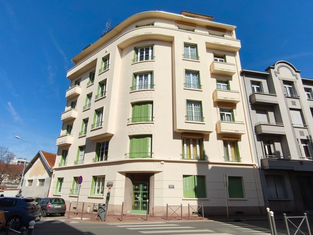 Un immeuble d'appartements de plusieurs étages, couleur crème, avec des volets verts, à un coin de rue sous un ciel bleu limpide.