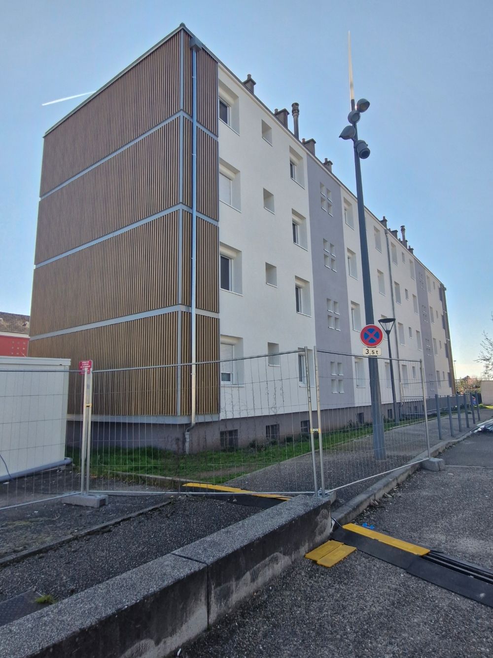 Un immeuble d'appartements à plusieurs étages avec un panneau latéral texturé moderne de couleur bronze, dissimulé derrière une clôture métallique temporaire.