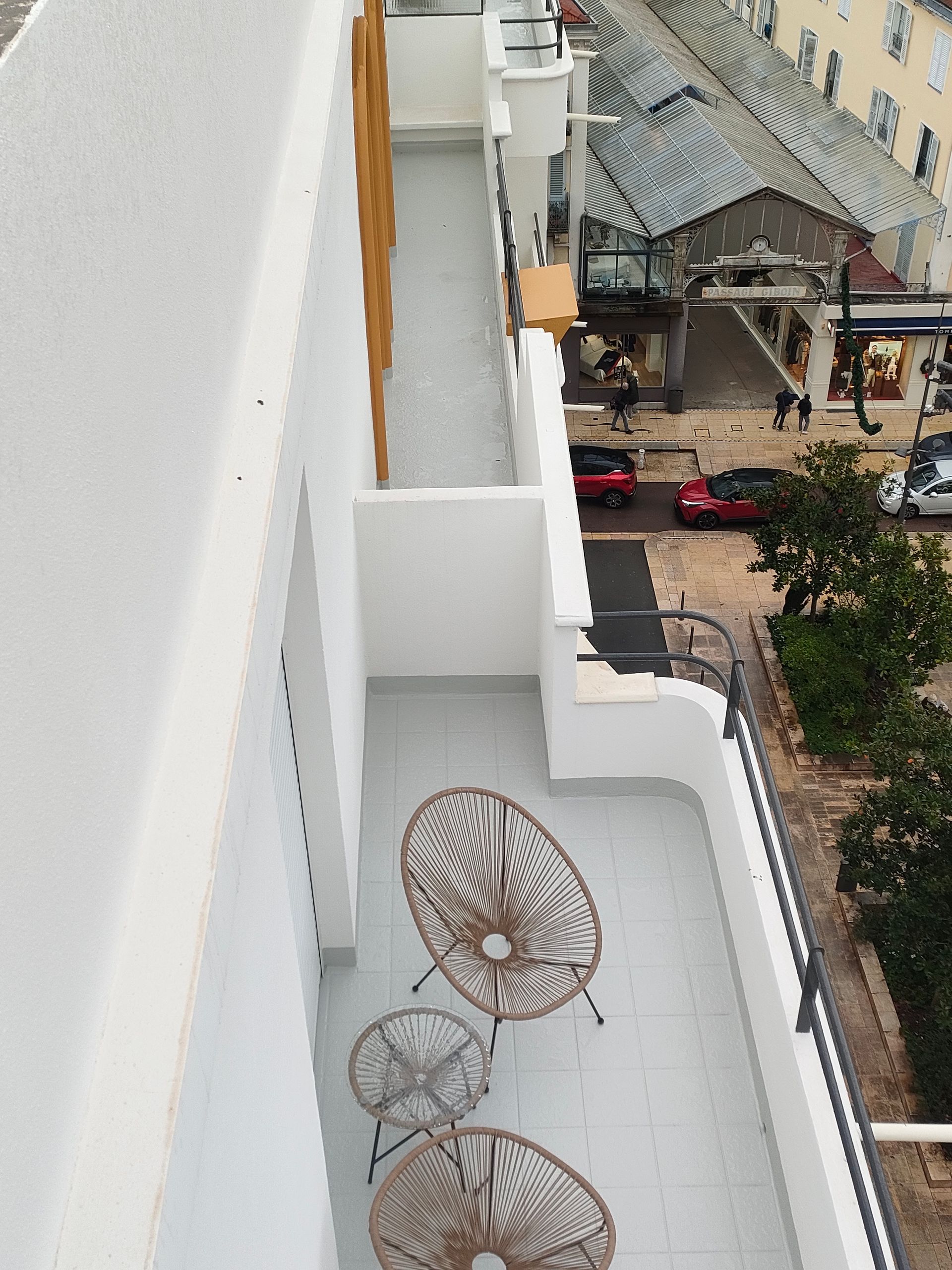 Vue en plongée d'un balcon blanc et moderne avec deux chaises en osier marron et une petite table, donnant sur une rue de la ville.