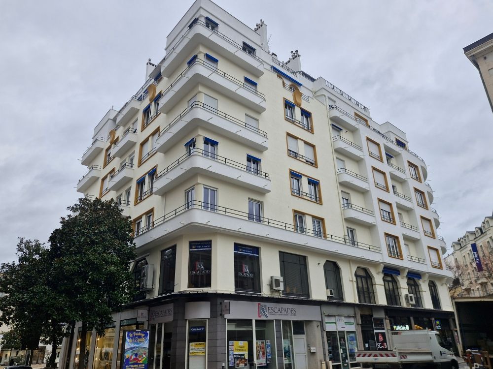 Un immeuble d'angle de cinq étages, de couleur crème, avec des balcons bleus et des devantures de magasins au rez-de-chaussée, sous un ciel couvert.