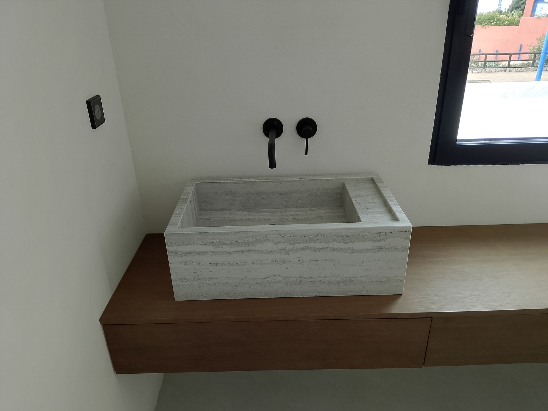 Lavabo rectangular de piedra sobre encimera de madera con grifería negra y ventana al fondo.