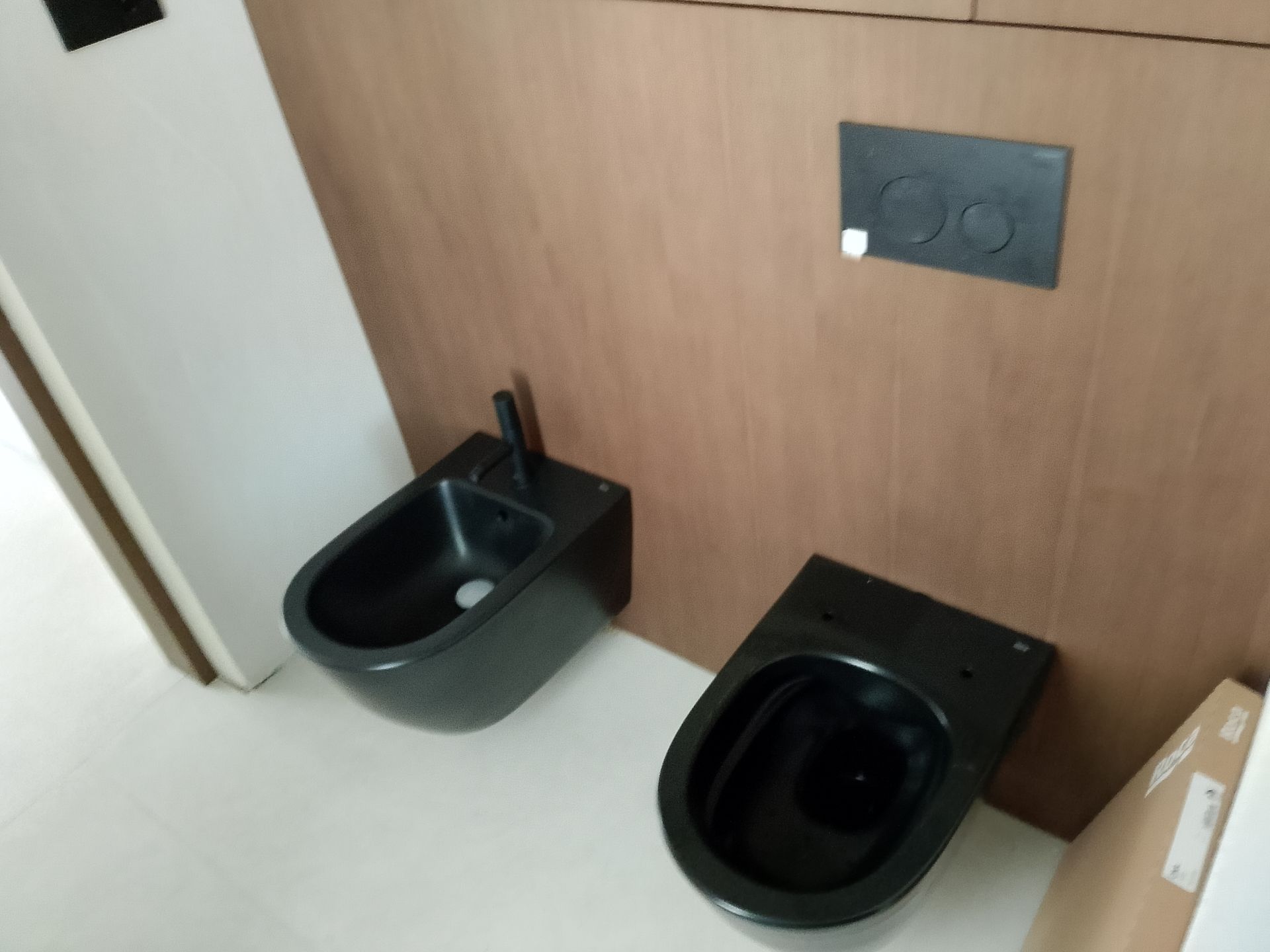 Inodoro y bidé negros montados en una pared revestida de madera, placa de descarga encima.