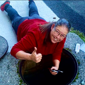 Eine Frau in rotem Hemd und Jeans liegt neben einem offenen Gullydeckel, zeigt den Daumen nach oben und lächelt. Sie hält ein Werkzeug in der Hand.