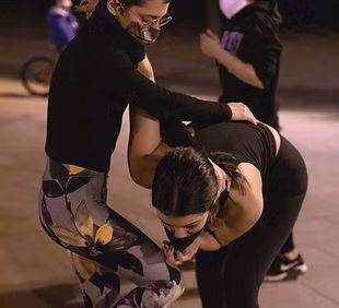 Dos mujeres están paradas una al lado de la otra en una acera. Una de las mujeres lleva una máscara.