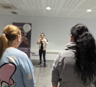 Un grupo de mujeres está de pie en una habitación mirando a un hombre que habla por un micrófono.