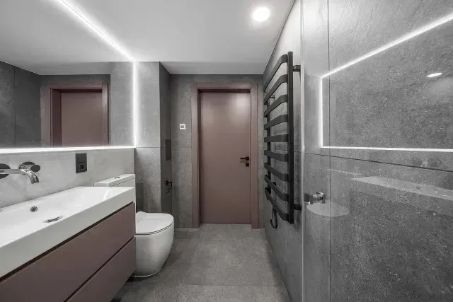 Baño moderno con azulejos grises, lavabo blanco, puerta rosa y calentador de toallas.