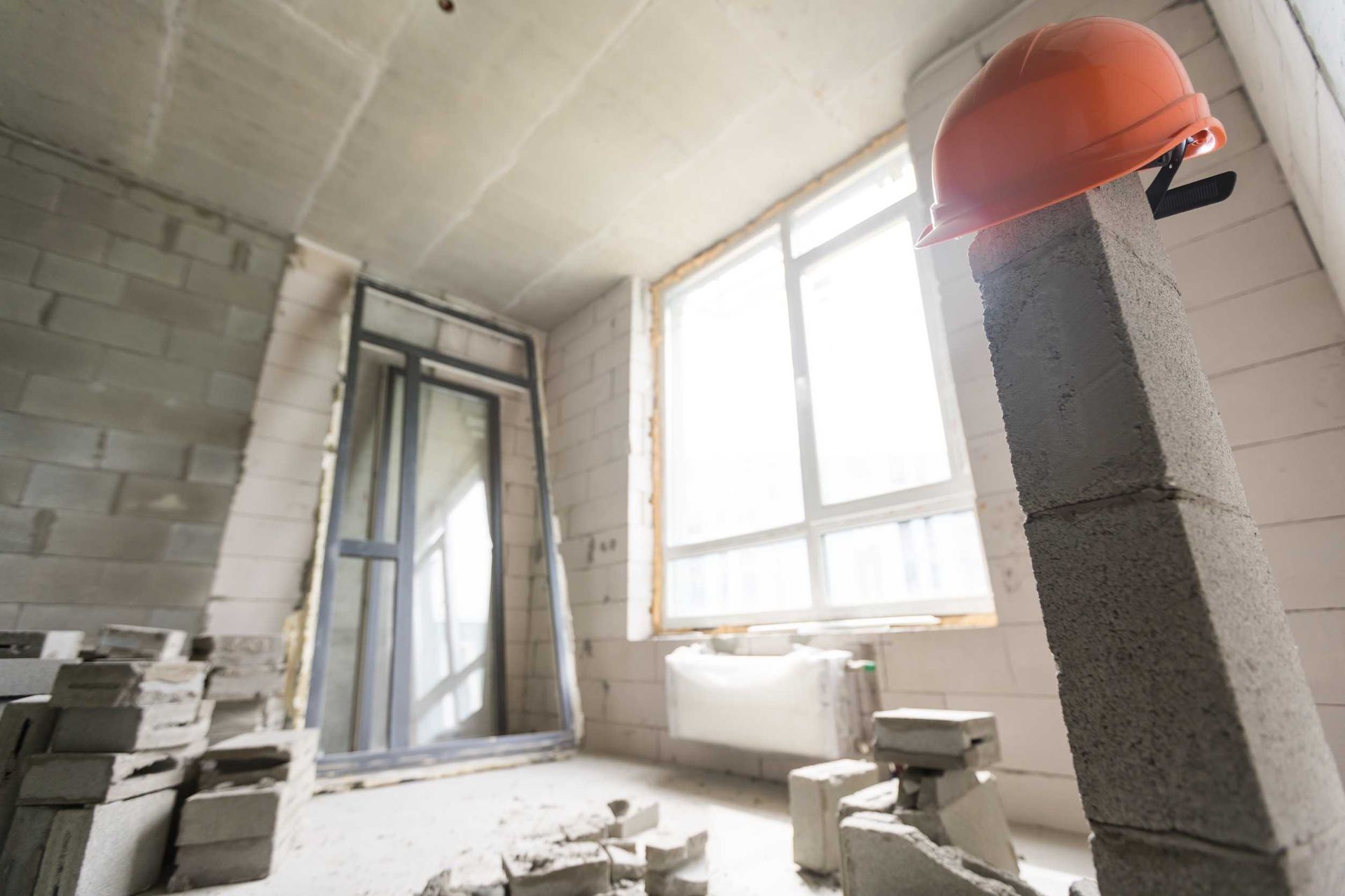 Casco naranja sobre un pilar de hormigón en una habitación en construcción, con una ventana y una puerta.