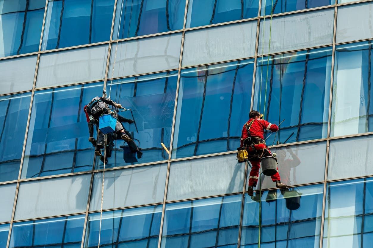 Dos limpiadores de ventanas limpiando el vidrio de un edificio alto; uno en azul, otro en rojo.