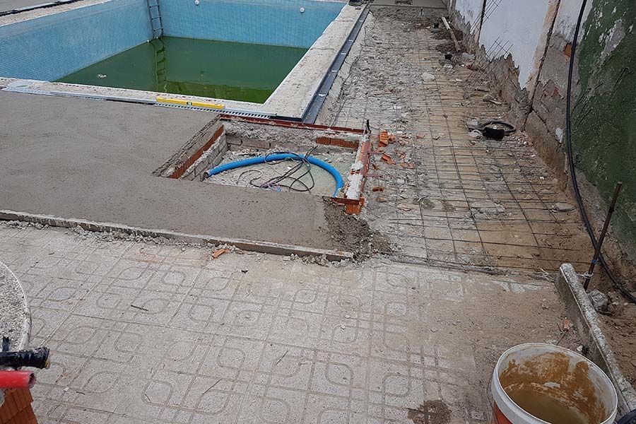 Renovación de la piscina en progreso: trabajo de hormigón alrededor de una piscina vacía, camino en demolición, plomería azul expuesta.