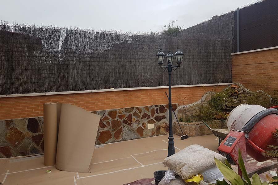 Patio trasero con cerca marrón, pared de ladrillo y farola. Hay materiales de construcción y una hormigonera.