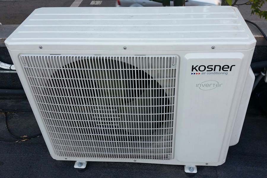 Unidad de aire acondicionado Kosner blanca sobre una superficie oscura; rejilla metálica con aspas de ventilador.