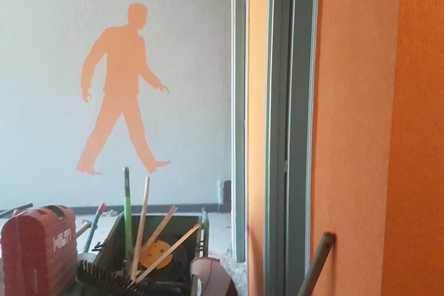 Silueta naranja de una persona caminando sobre un muro. Hay herramientas y una carretilla frente al muro.