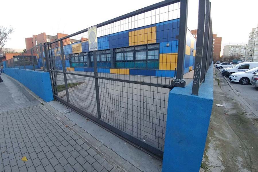 Exterior de un edificio azul, amarillo y verde azulado tras una valla metálica y un muro azul. Coches aparcados a la derecha.