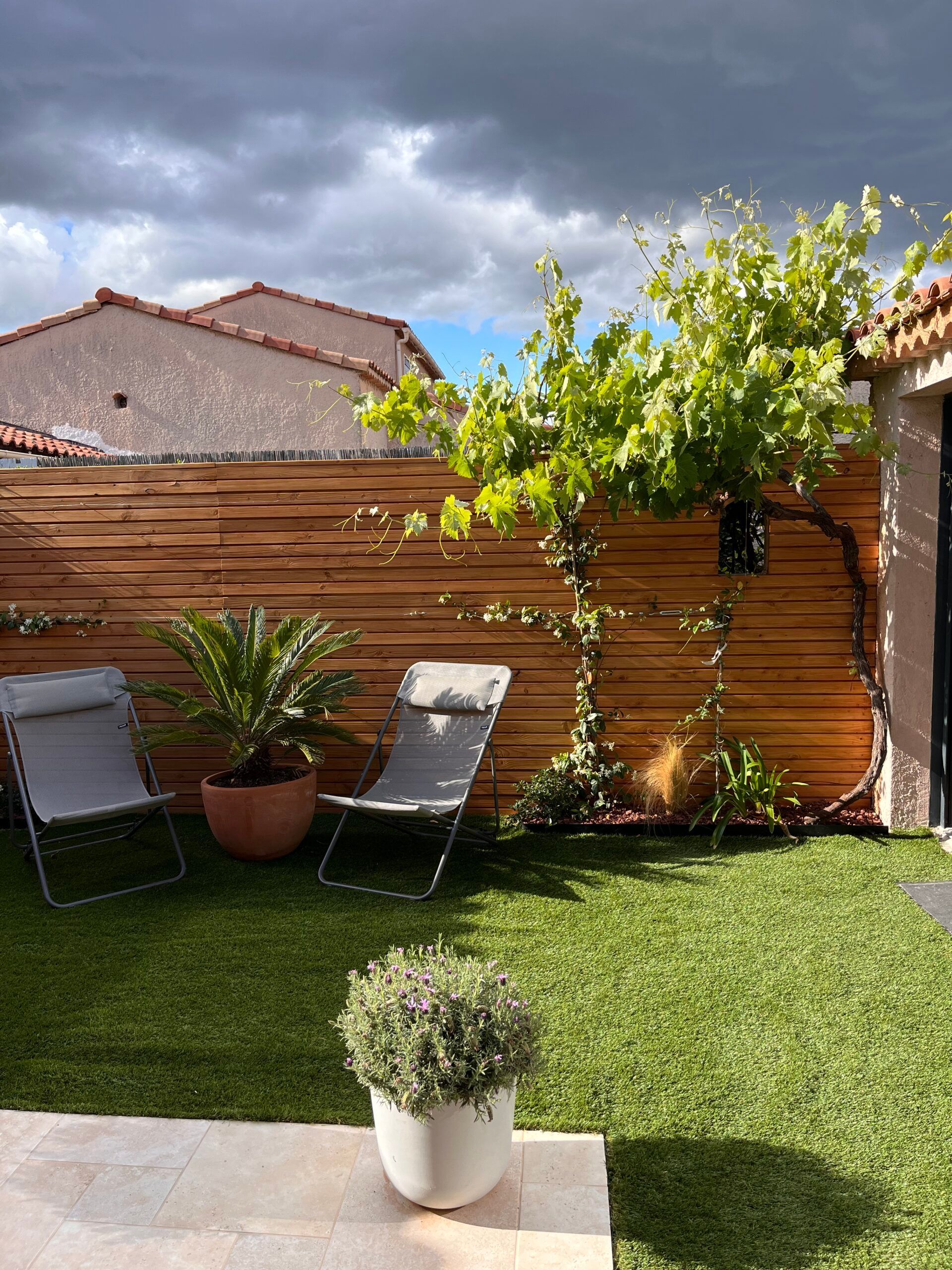 Une cour arrière avec une clôture en bois, des chaises, des plantes en pot et un arbre.
