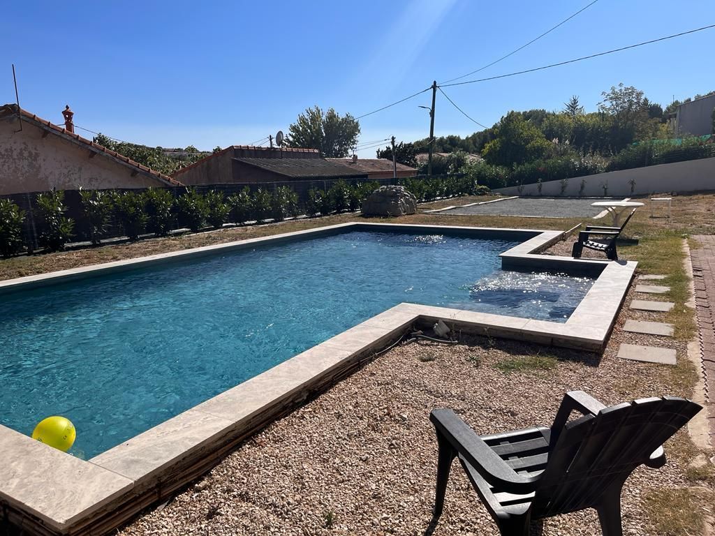 Piscine avec eau bleu clair, patio en pierre, chaise, cour en gravier et arbres sous un ciel ensoleillé.