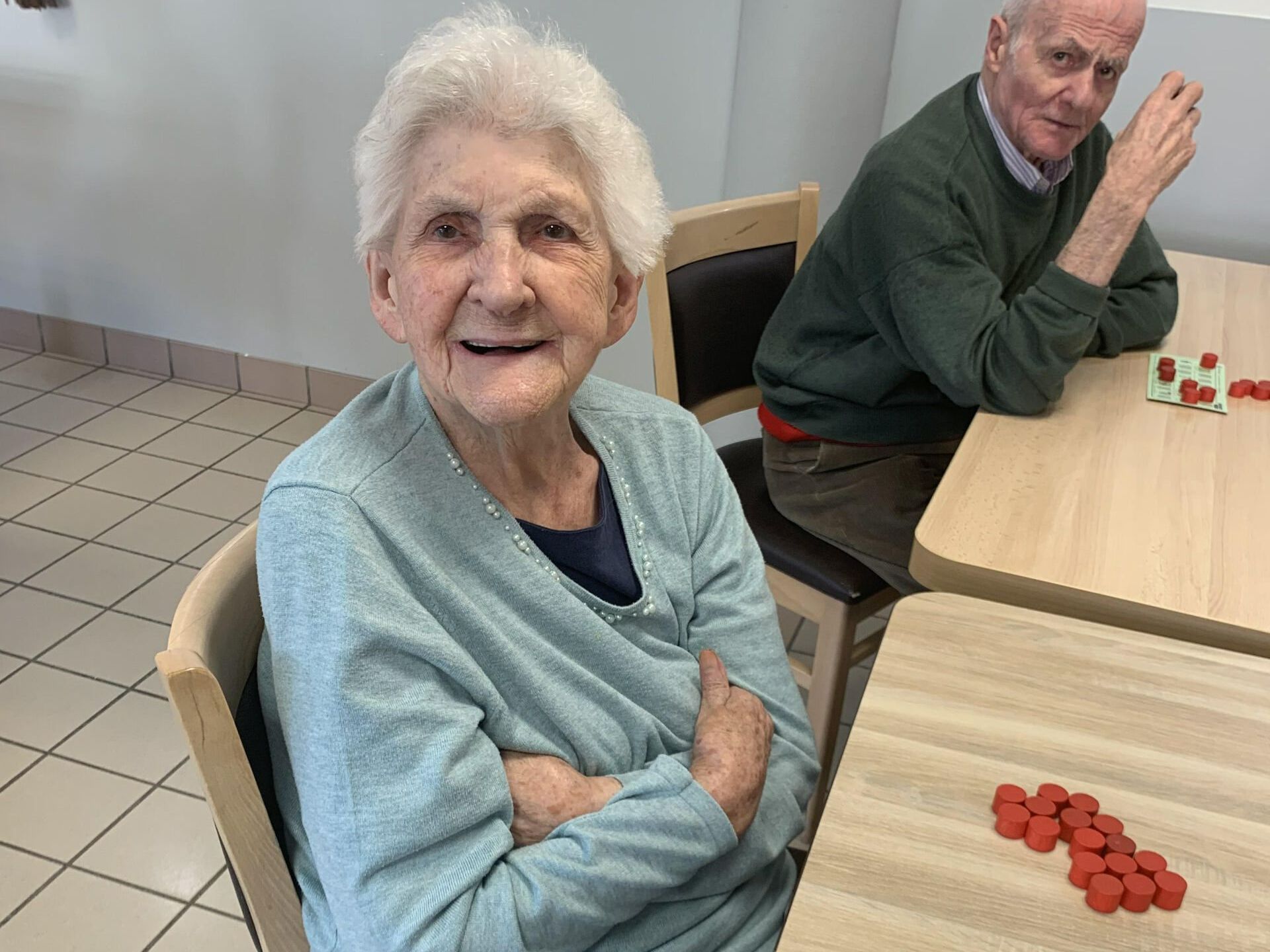 Une femme âgée sourit, les bras croisés, avec un homme en arrière-plan à une table jouant à un jeu avec des pièces rouges.