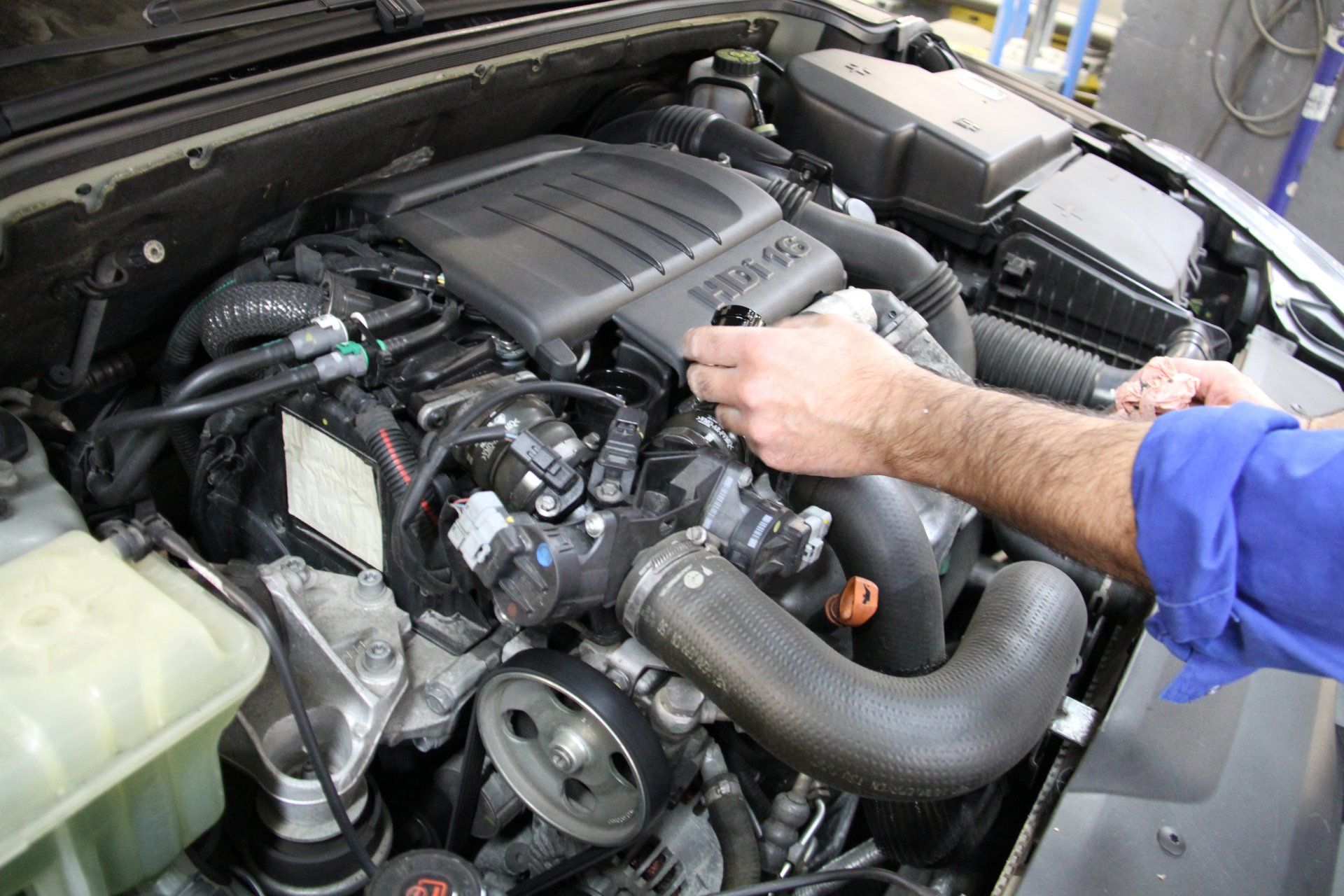 Des mains travaillent sur le moteur d'une voiture dans le compartiment moteur. On aperçoit le cache-moteur noir et divers tuyaux.