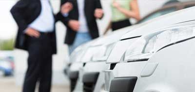 Des personnes examinent des voitures chez un concessionnaire. Des voitures blanches sont alignées, avec des personnes en costume en arrière-plan.