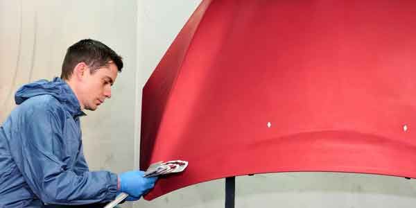 Une personne pulvérise de la peinture rouge sur le capot d'une voiture. Elle porte une veste bleue et des gants dans un atelier.