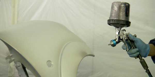 Une personne applique de la peinture au pistolet sur une pièce automobile dans un atelier. La pièce est gris clair.