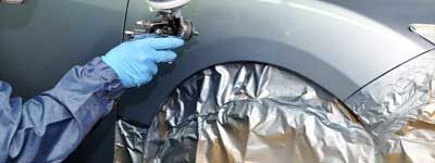 Personne peignant une voiture au pistolet à peinture, portant des vêtements de protection et des gants.