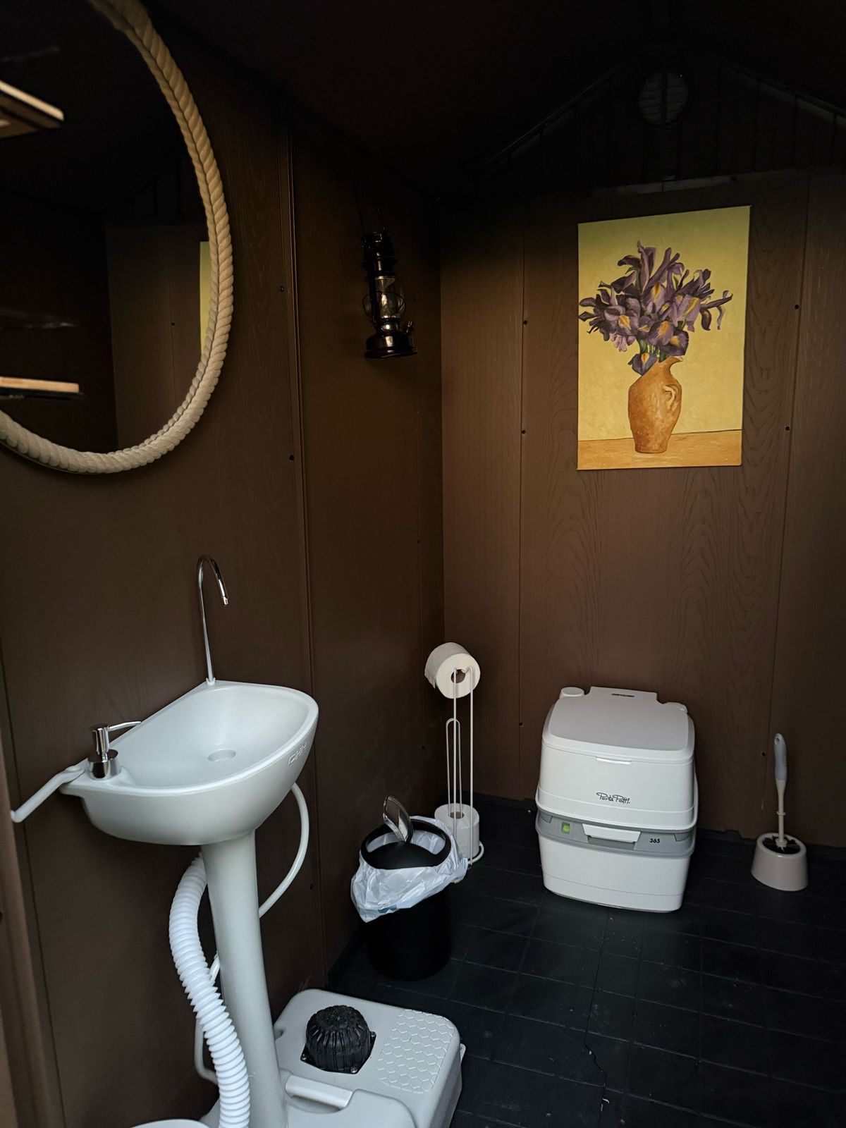 Baño pequeño de color marrón oscuro con lavabo, inodoro portátil, obras de arte y espejo. Papelera negra, portarrollos y escobilla.