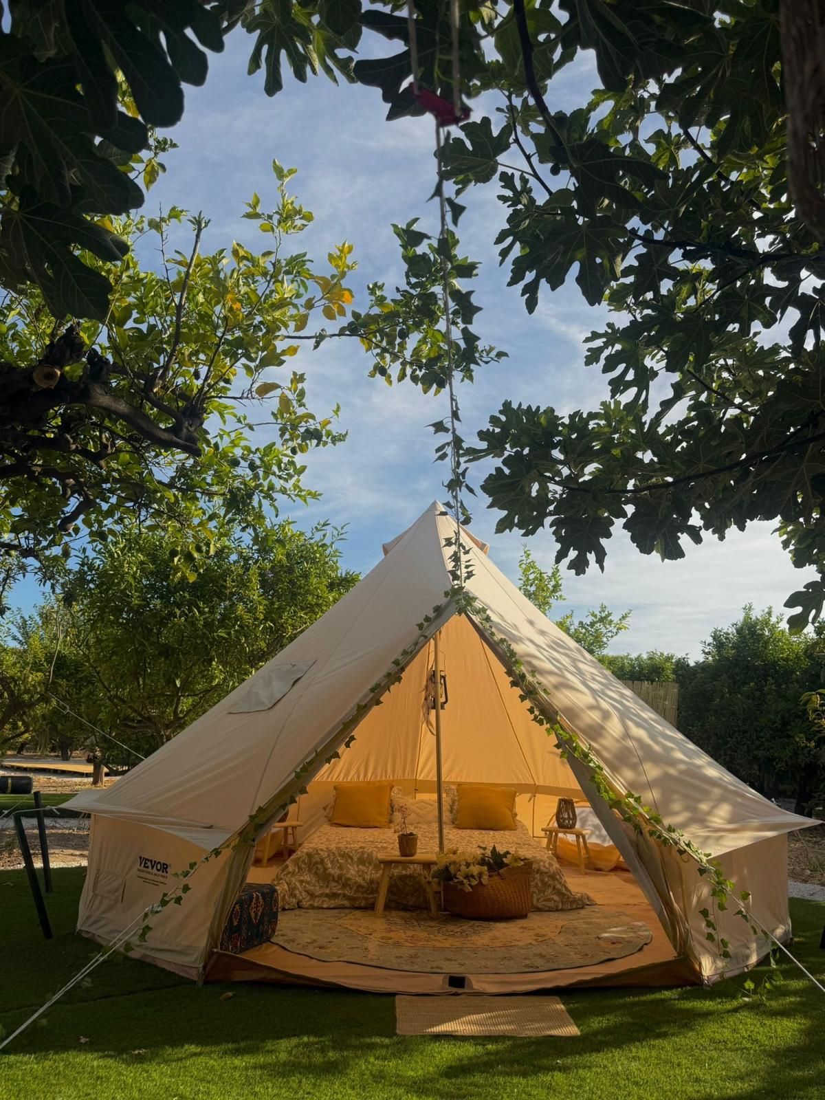 Tienda de campaña de color beige, abierta, decorada con almohadas, situada sobre el césped, rodeada de árboles bajo un cielo azul.