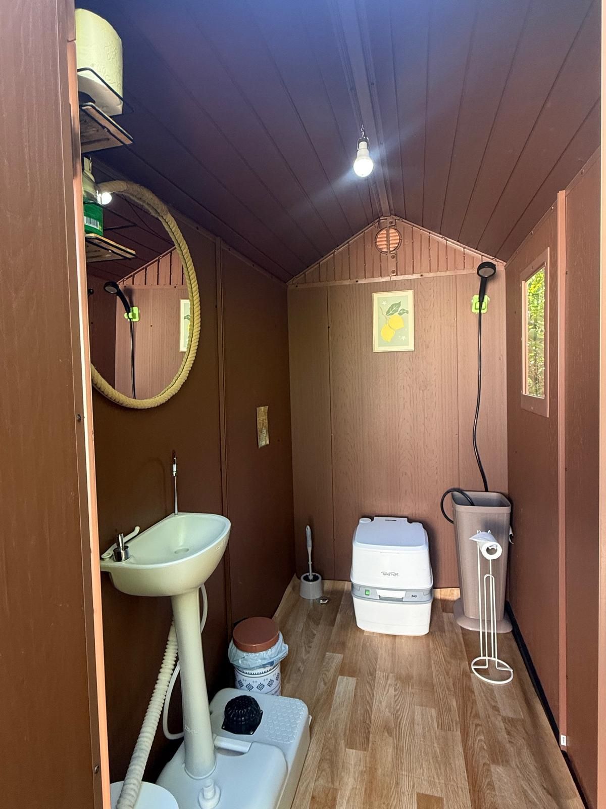 Baño pequeño, rústico, de paredes marrones, con lavabo, inodoro de compostaje, cabezal de ducha, espejo y luz.