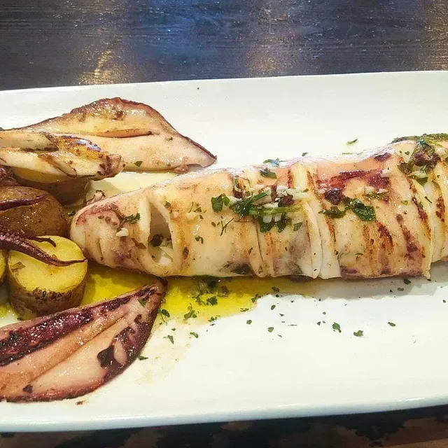 Calamares a la plancha en un plato blanco, servidos con patatas y rociados con aceite de oliva y hierbas.