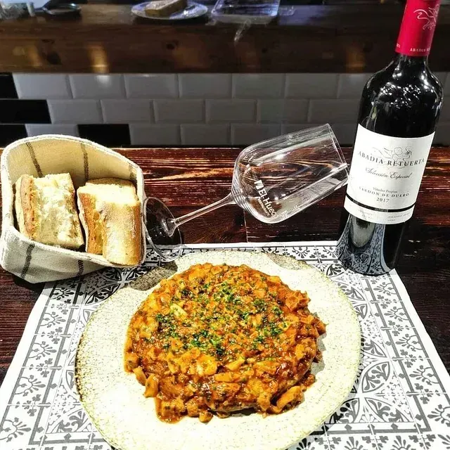 Un plato de frijoles guisados, pan, una botella de vino y una copa sobre una mesa de un bar.