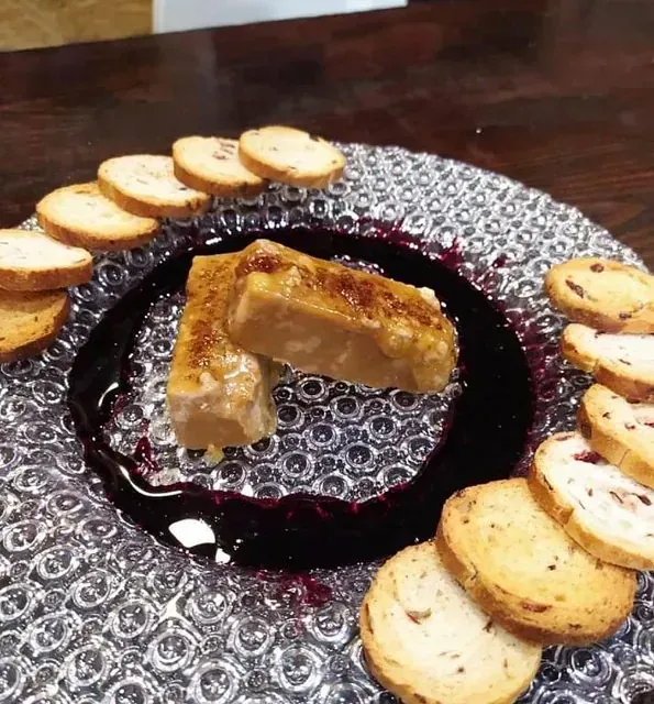 Dos aperitivos rectangulares con superficies caramelizadas sobre un plato transparente con un aro de salsa oscura y rodajas de pan tostado.