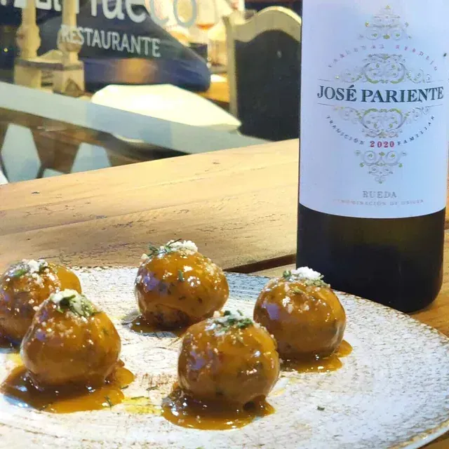 Albóndigas con salsa en plato, acompañadas de una botella de vino José Pariente Rueda, con un ambiente de restaurante de fondo.