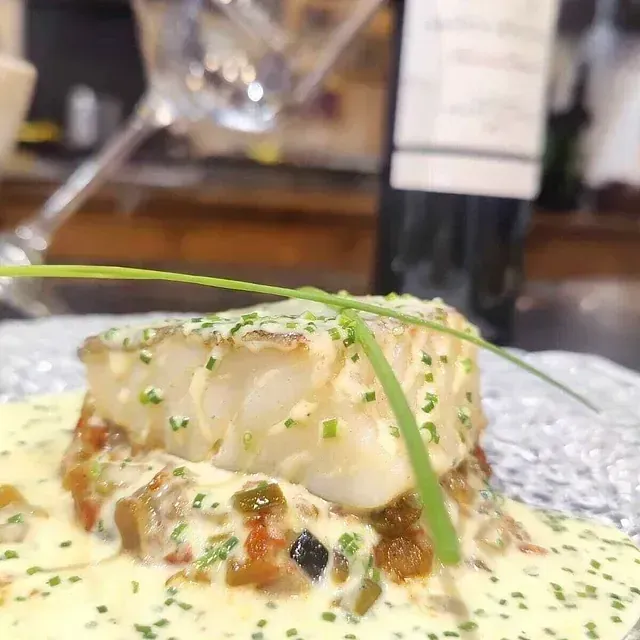Filete de bacalao a la plancha con salsa cremosa, verduras y guarnición de cebollino, servido en un plato decorativo. Copa de vino y botella al fondo.