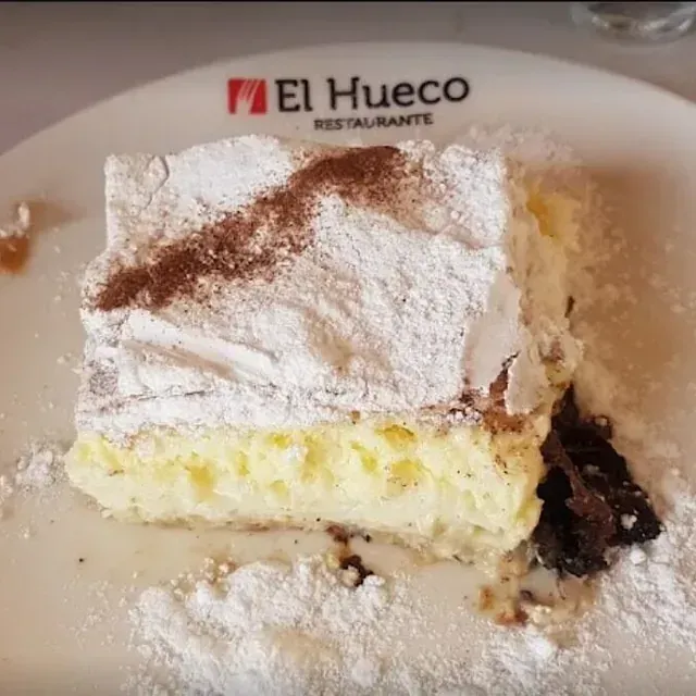 Una rebanada de postre de hojaldre en capas espolvoreado con azúcar en polvo y cacao, servido en un plato blanco en el restaurante El Hueco.
