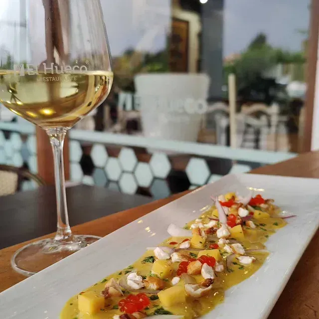 Una copa de vino blanco junto a un plato rectangular con salsa amarilla, mango y nueces, con un restaurante al fondo.