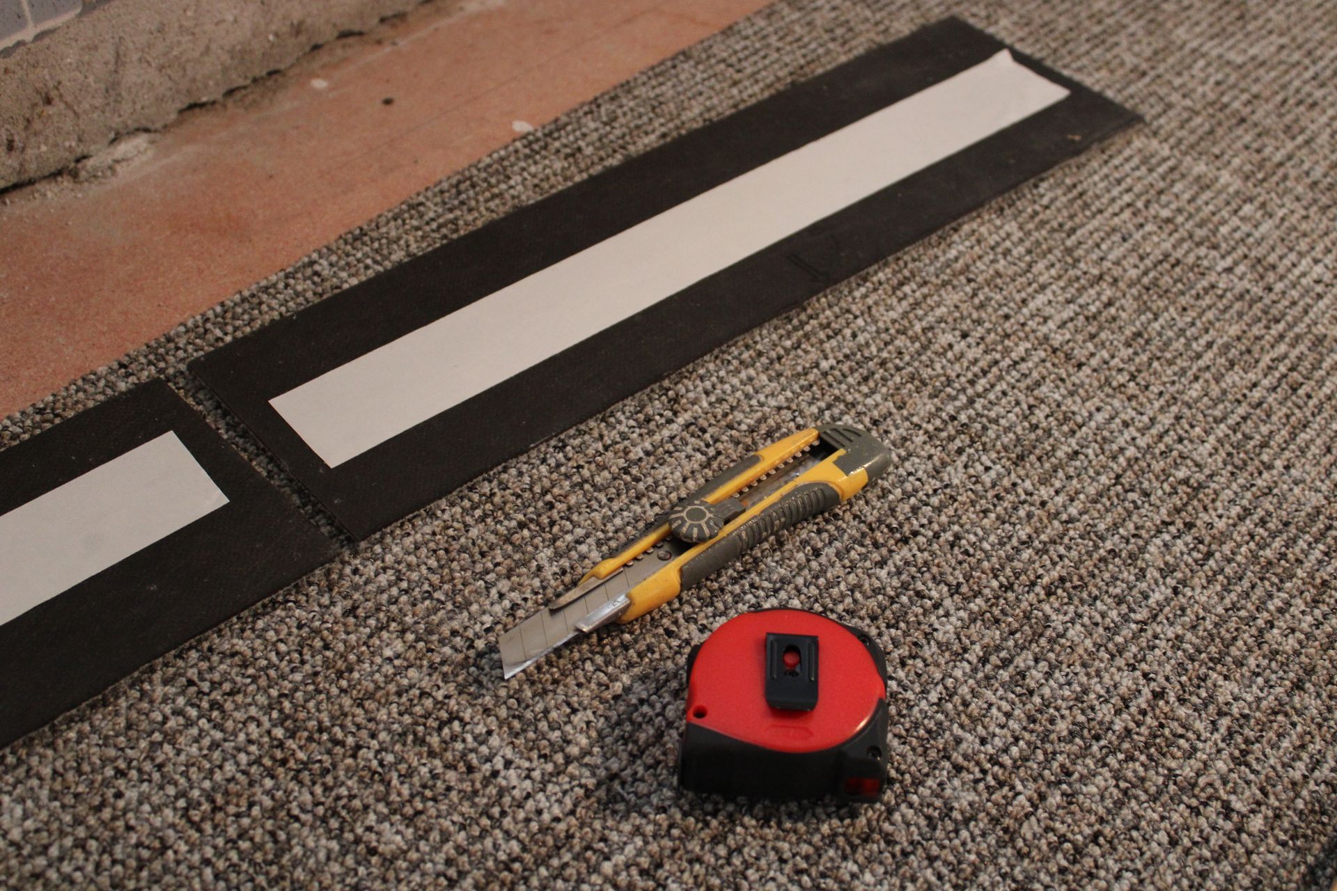 Dalles de moquette, cutter et mètre ruban sur un sol recouvert de moquette à motifs.