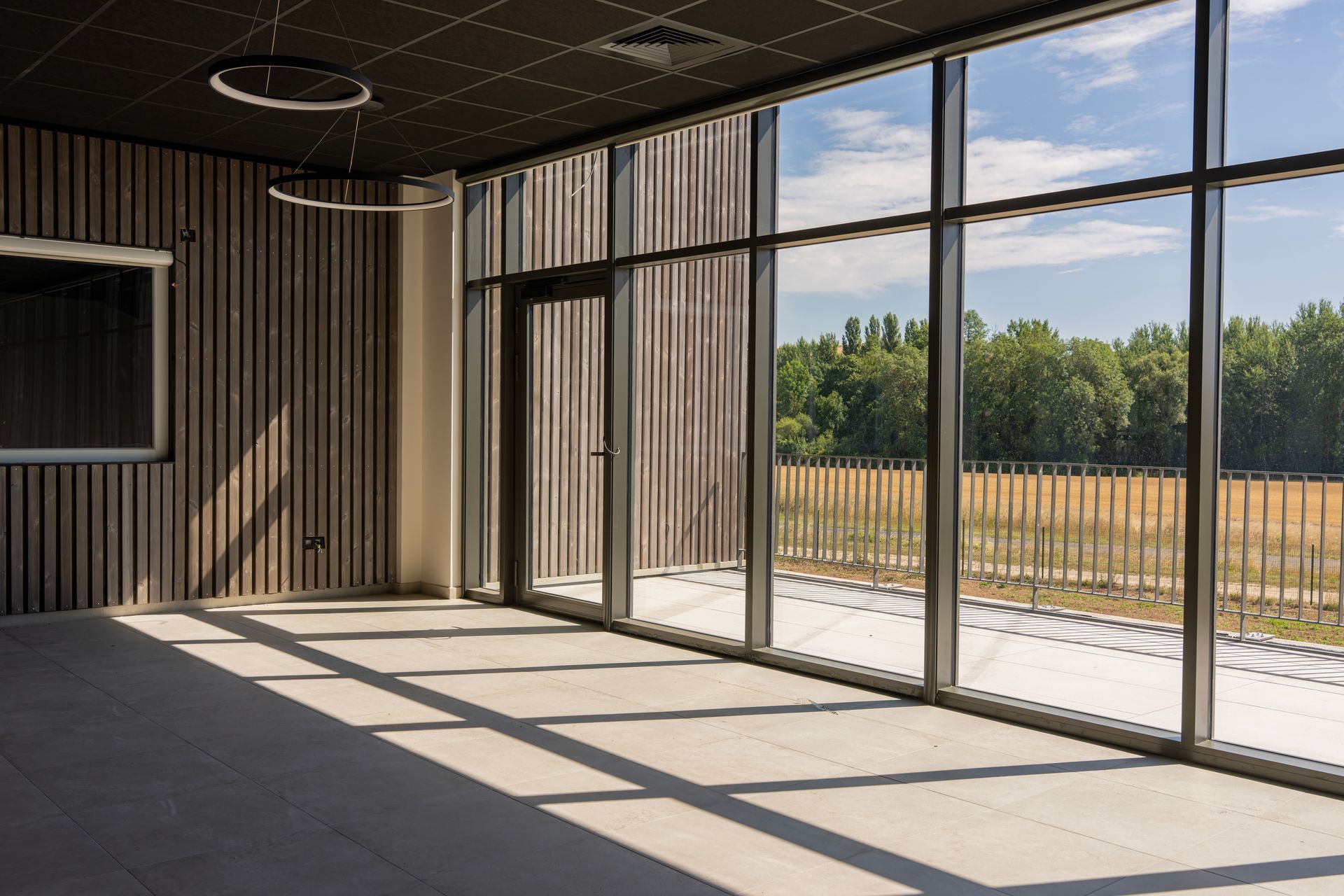 Vue intérieure avec de grandes fenêtres, montrant une pièce lumineuse et une vue sur des arbres et un champ.