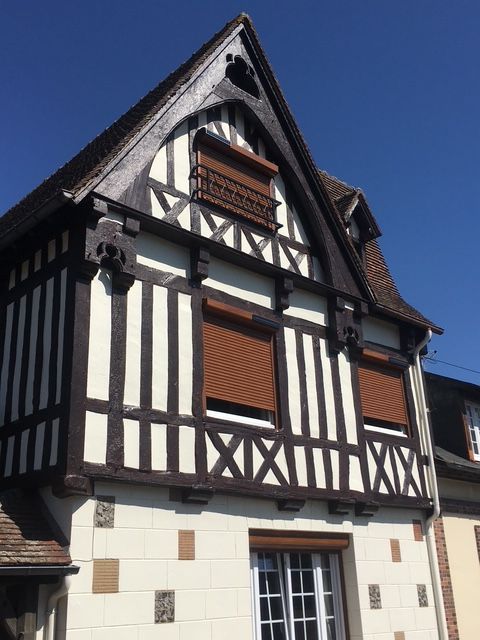 Maison à colombages avec poutres brunes, murs blancs, volets bruns et ciel bleu.
