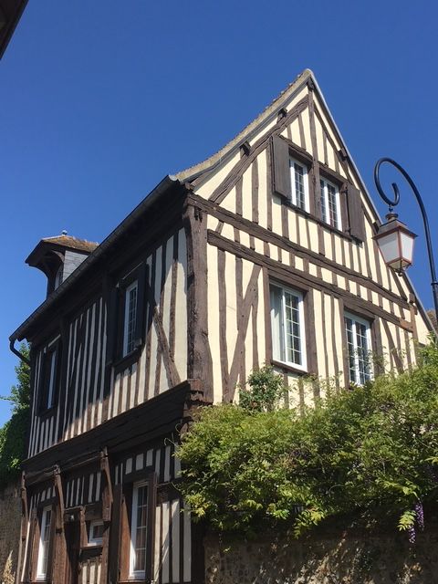 Bâtiment avec colombages de deux étages, avec panneaux blancs, poutres brunes et volets, sous un ciel bleu.