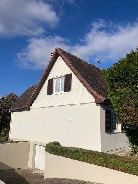 Bâtiment de style cottage avec bardage blanc, toit et volets bruns, une haie et un ciel bleu nuageux.