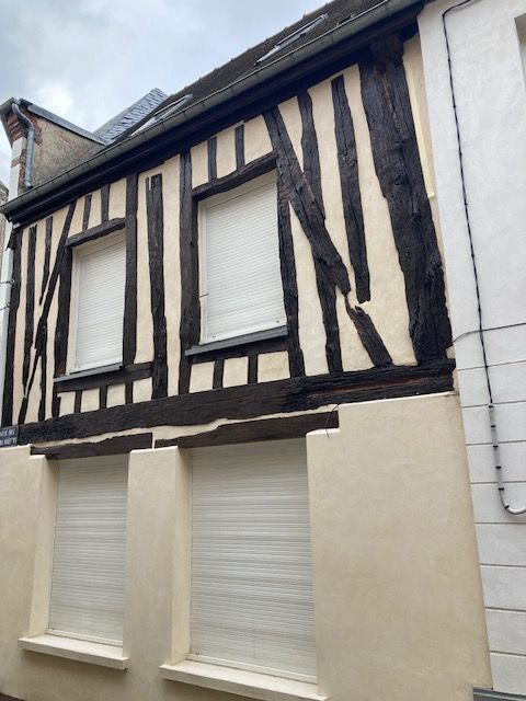 Bâtiment à colombages avec poutres en bois sombre et remplissage clair. Deux fenêtres à volets à chaque étage.