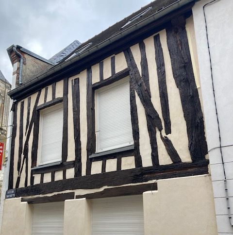 Bâtiment à colombages avec poutres noires et remplissage couleur crème. Deux fenêtres à stores blancs sont visibles.