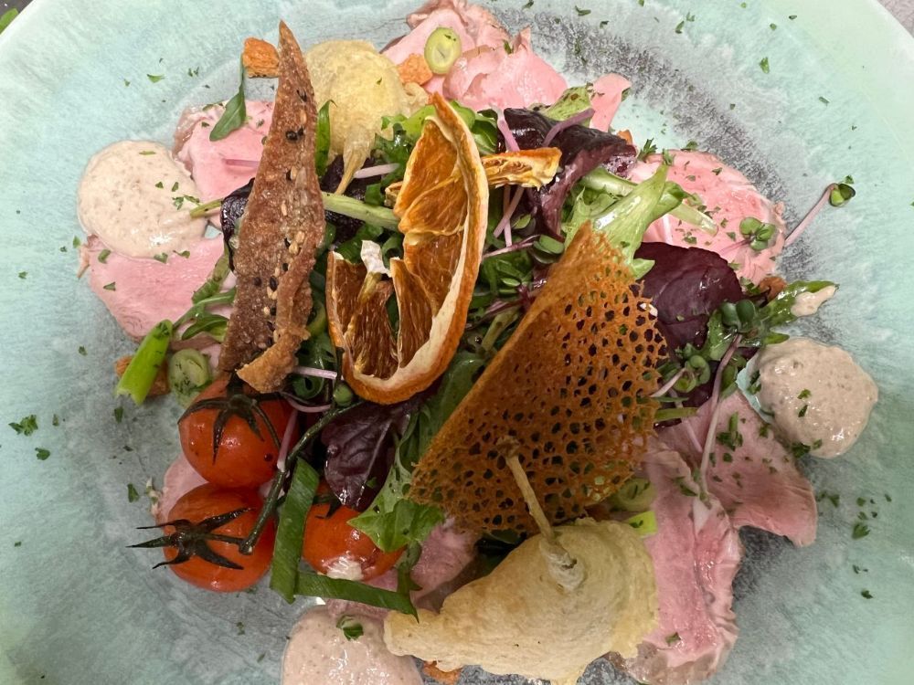 Salat mit geschnittenem Fleisch, Kirschtomaten, Orangenscheibe und knusprigem Brot auf einem blauen Teller.