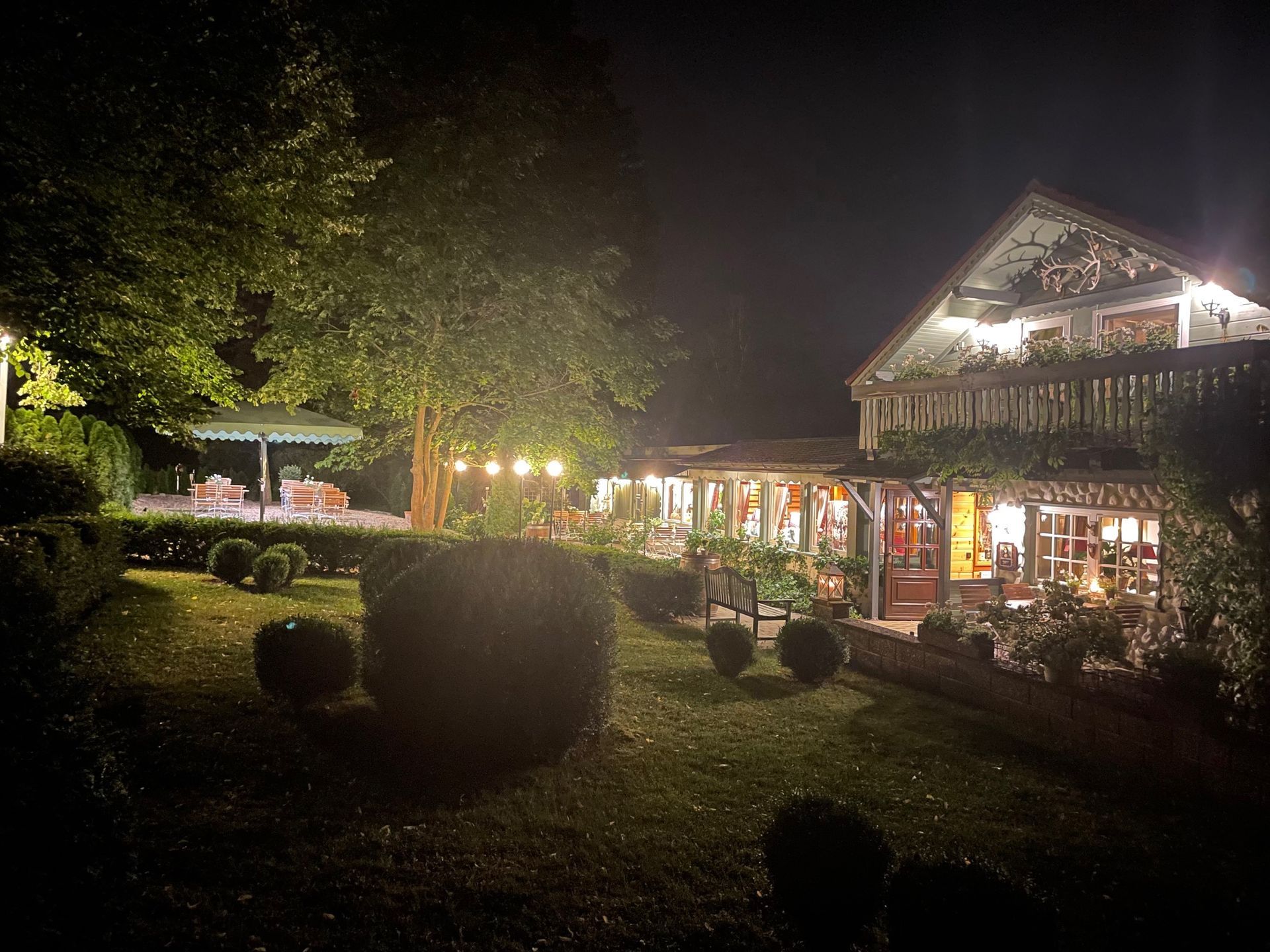 Nachts beleuchteter Außenbereich des Restaurants; Garten mit gestutzten Büschen, Bäumen und Sitzgelegenheiten im Freien; warmes Licht.