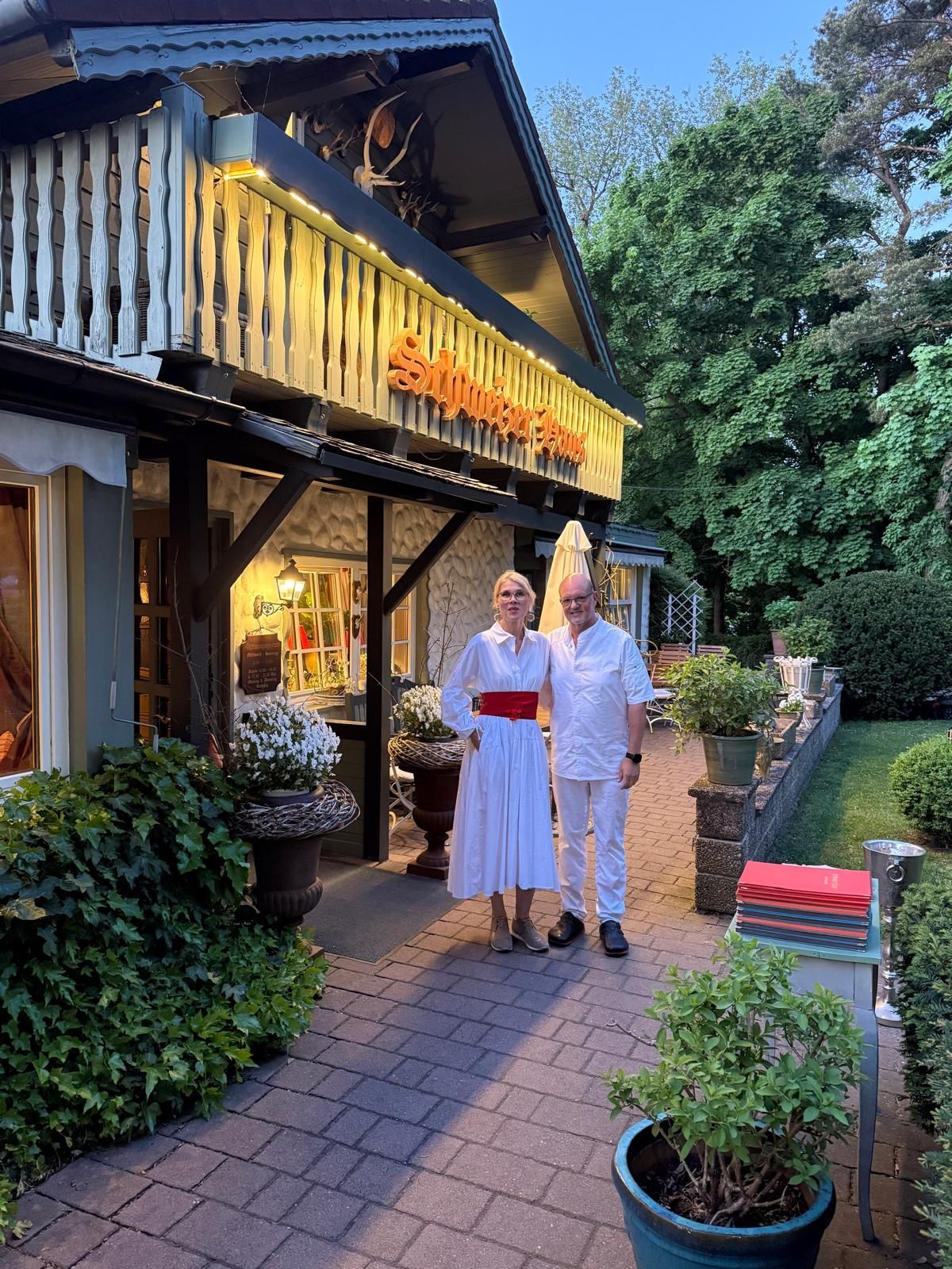 Paar steht vor einem Restaurant mit Schild. Frau trägt weißes Kleid mit roter Schärpe. Mann im weißen Anzug.