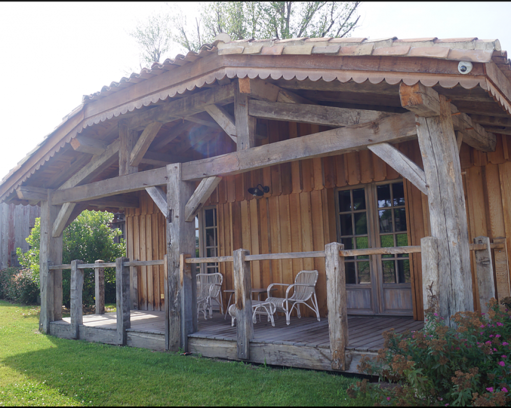 Véranda rustique en bois avec poutres apparentes, fauteuils en osier et petite table, à côté d’une cabane en rondins dans un jardin herbeux