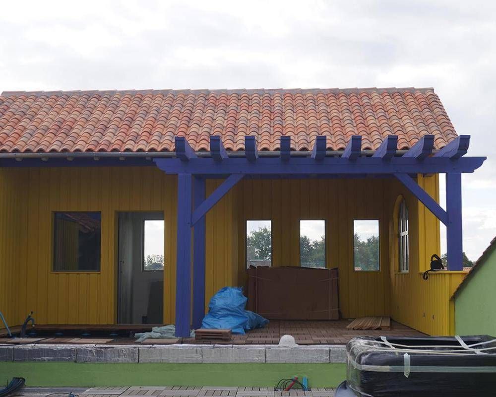Maison jaune avec des poutres de porche bleues et un toit de tuiles, avec un tas recouvert d’une bâche bleue sur le porche.